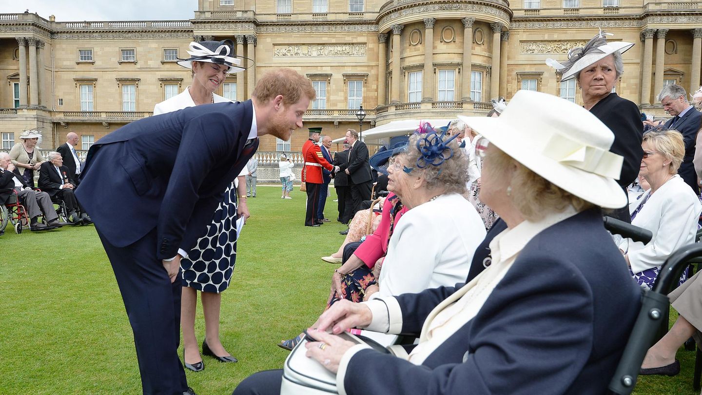 Prinz Harry im dunklen Anzug beugt sich zu älteren Damen im Rollstuhl herunter, mit denen er plaudert