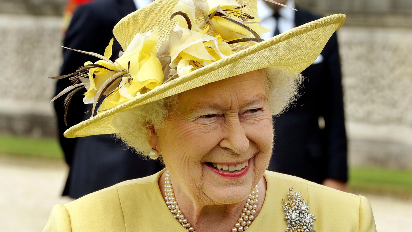 Die Queen trägt einen große runden gelben Hut und lächelt