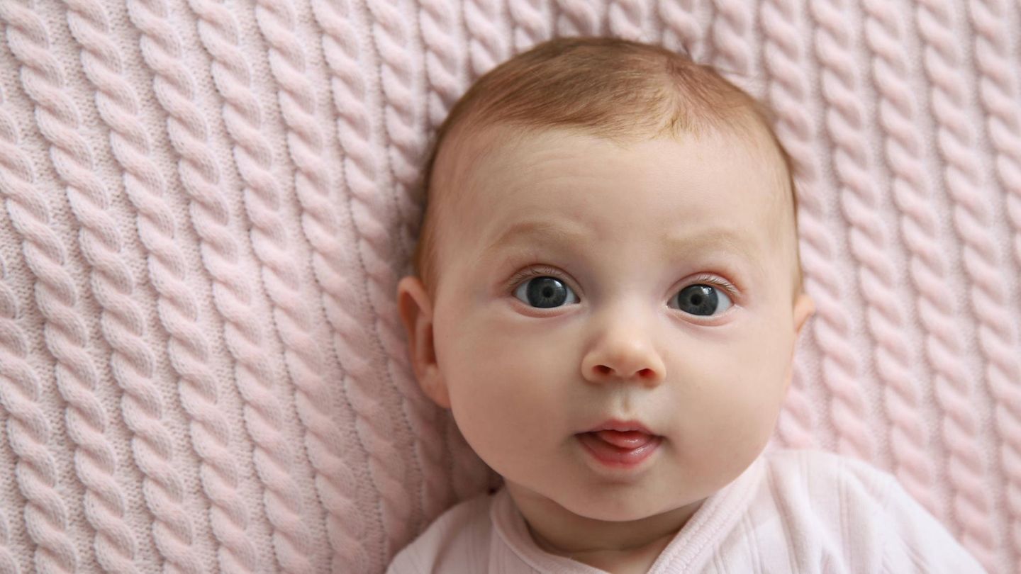 Babynamen: Ein weißes Baby liegt auf einer rosa Strickdecke und schaut direkt in die Kamera