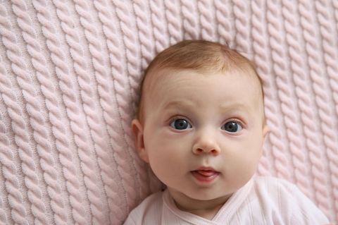 Babynamen: Ein weißes Baby liegt auf einer rosa Strickdecke und schaut direkt in die Kamera
