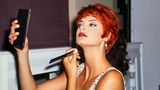 Linda Evangelista mit kurzen roten Haaren blickt in einen Spiegel und pudert sich das Kinn
