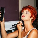Linda Evangelista mit kurzen roten Haaren blickt in einen Spiegel und pudert sich das Kinn