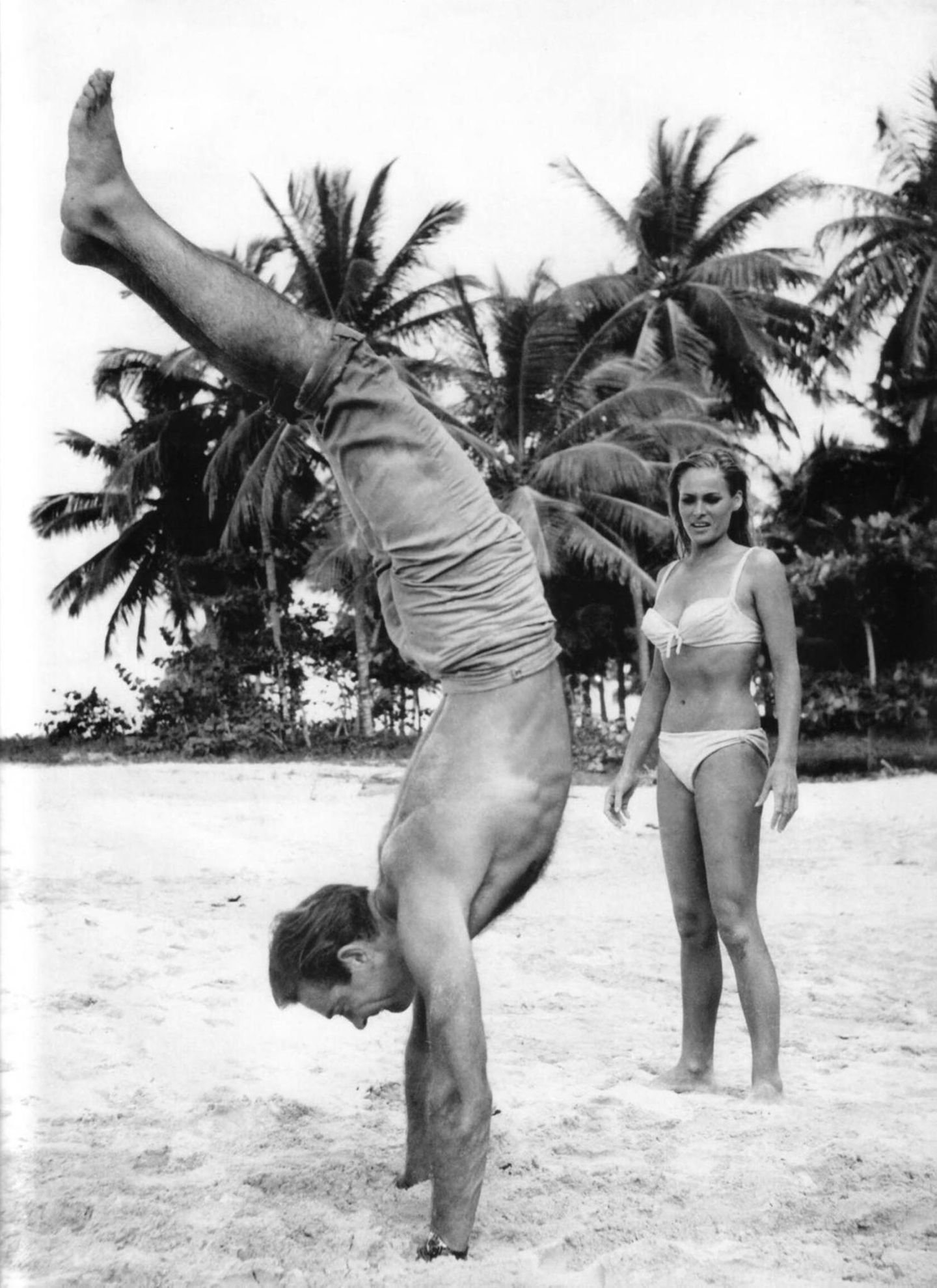 Sean Connery macht Handstand am Strand, im Hintergrund steht Ursula Andress im Bikini