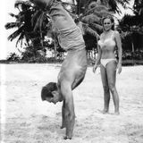 Sean Connery macht Handstand am Strand, im Hintergrund steht Ursula Andress im Bikini