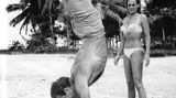 Sean Connery macht Handstand am Strand, im Hintergrund steht Ursula Andress im Bikini