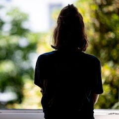 Der Umriss einer jungen Frau vor einem offenen Fenster. Draußen, verschwommen im Hintergrund sind Bäume und Sonnenschein.