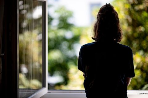 Der Umriss einer jungen Frau vor einem offenen Fenster. Draußen, verschwommen im Hintergrund sind Bäume und Sonnenschein.
