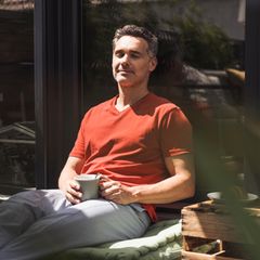 Ein Mann sitzt in der Sonne mit geschlossenen Augen und einer Tasse in der Hand