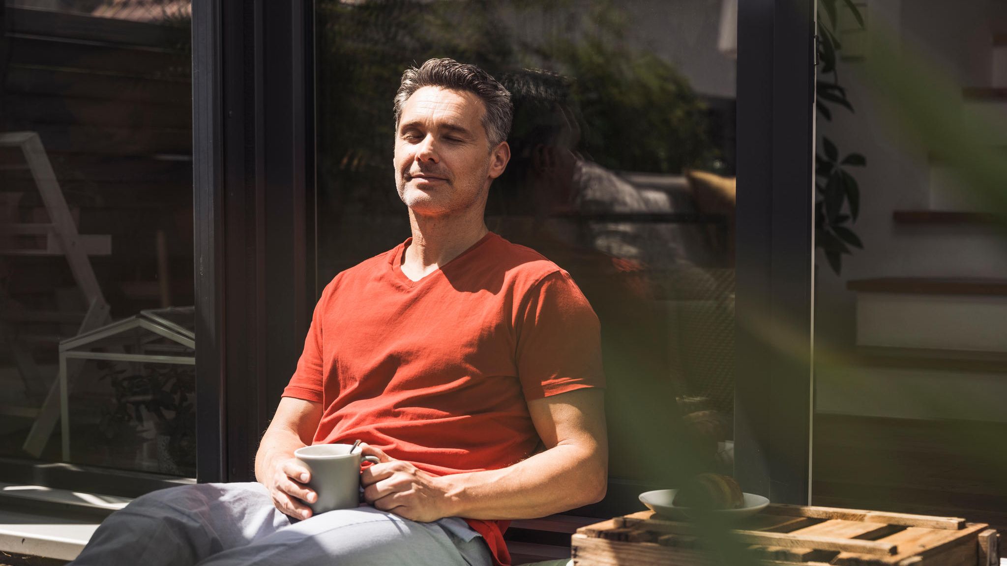 Ein Mann sitzt in der Sonne mit geschlossenen Augen und einer Tasse in der Hand