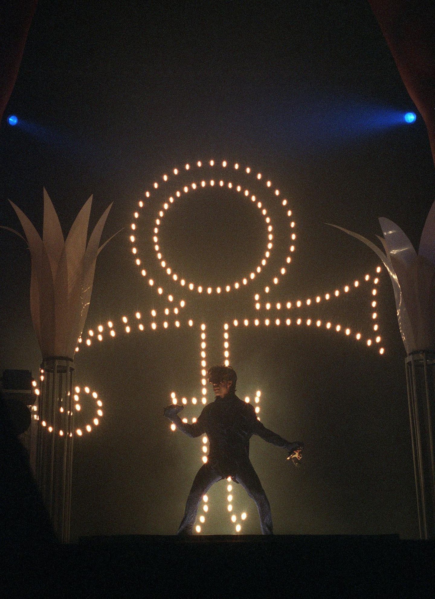 Prince im Dunkel der Bühne vor einem leuchtenden Symbol