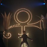 Prince im Dunkel der Bühne vor einem leuchtenden Symbol
