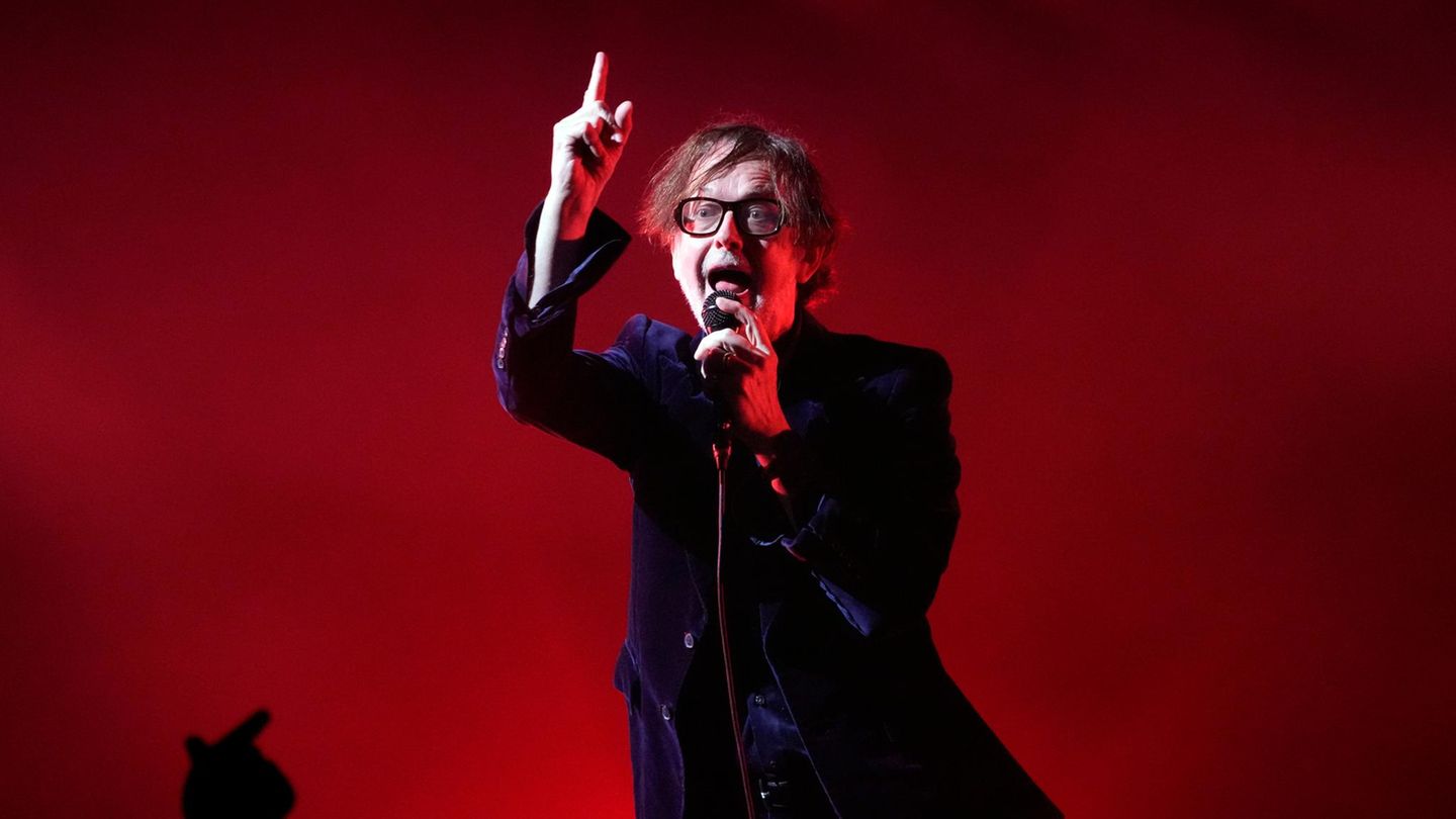 Der Sänger der Band Pulp, Jarvis Cocker, singend vor rotem Hintergrund. Er trägt ein dunkelblaues Samtjackett
