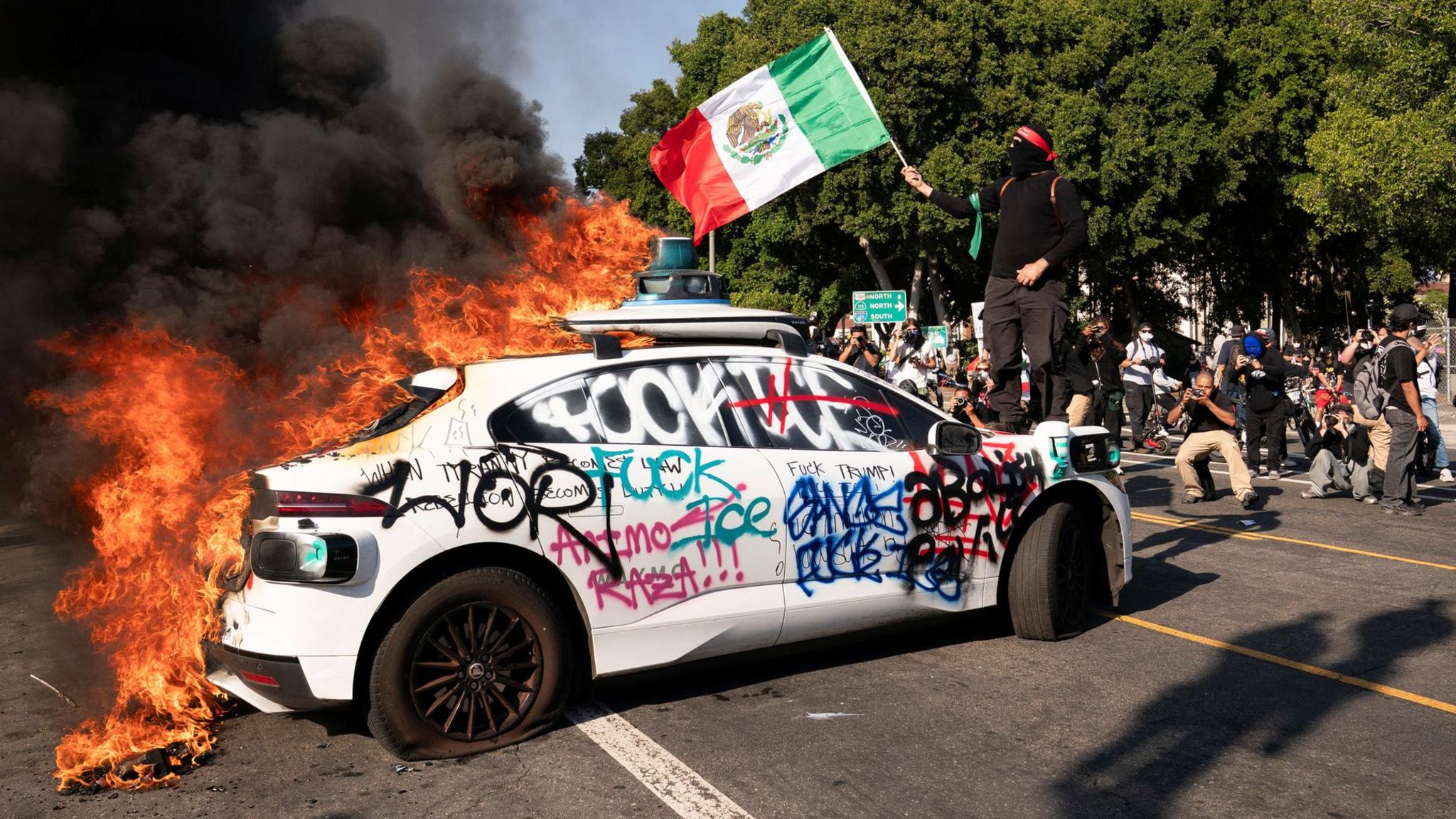 Szenen aus Los Angeles: Ein vermummter Demonstrant steht mit Mexiko-Flagge auf der Motorhaube eines brennenden Autos, auf das "Fuck ICE" geschmiert wurde. ICE ist die Einwanderungsbehörde der USA, die großflächig im Land auf Anweisung Trumps Razzien gegen illegale Einwanderer durchführt