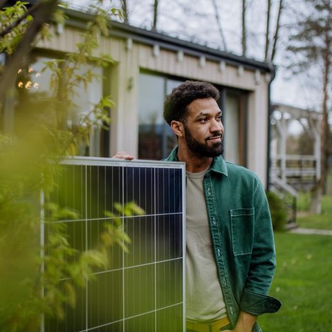 A man with the solar panel of a balcony power plant in front of a house framed by green leaves of a tree.
