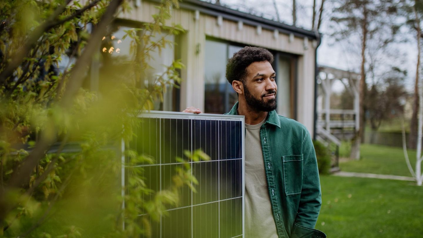 Ein Mann mit dem Solarpanel eines Balkonkraftwerkes vor einem Haus, das von Grünen Blättern eines Baumes eingerahmt ist.