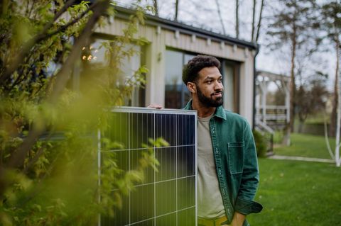 Ein Mann mit dem Solarpanel eines Balkonkraftwerkes vor einem Haus, das von Grünen Blättern eines Baumes eingerahmt ist.
