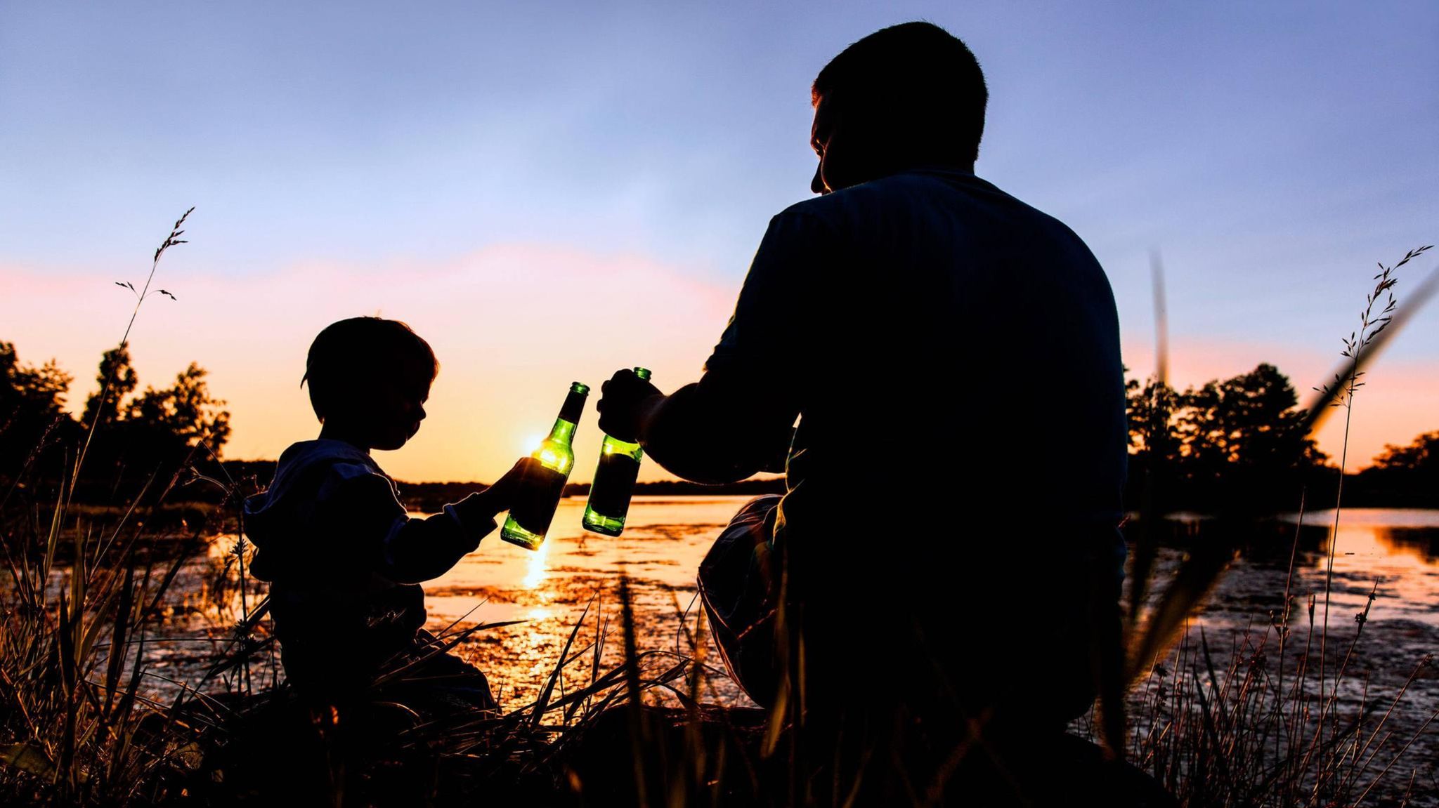 Erziehung: Vater und Sohn sitzen bei Sonnenuntergang am Wasser und stoßen mit Flaschen an