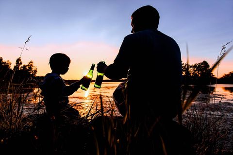 Erziehung: Vater und Sohn sitzen bei Sonnenuntergang am Wasser und stoßen mit Flaschen an