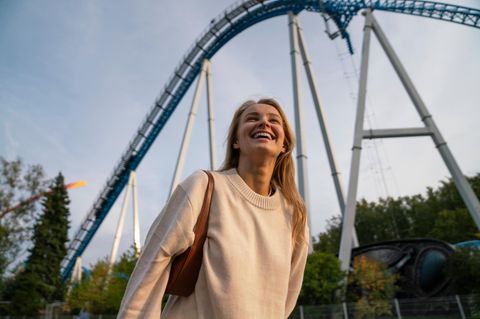 Vor einer Achterbahn steht eine junge Frau im hellen Pullover und lacht
