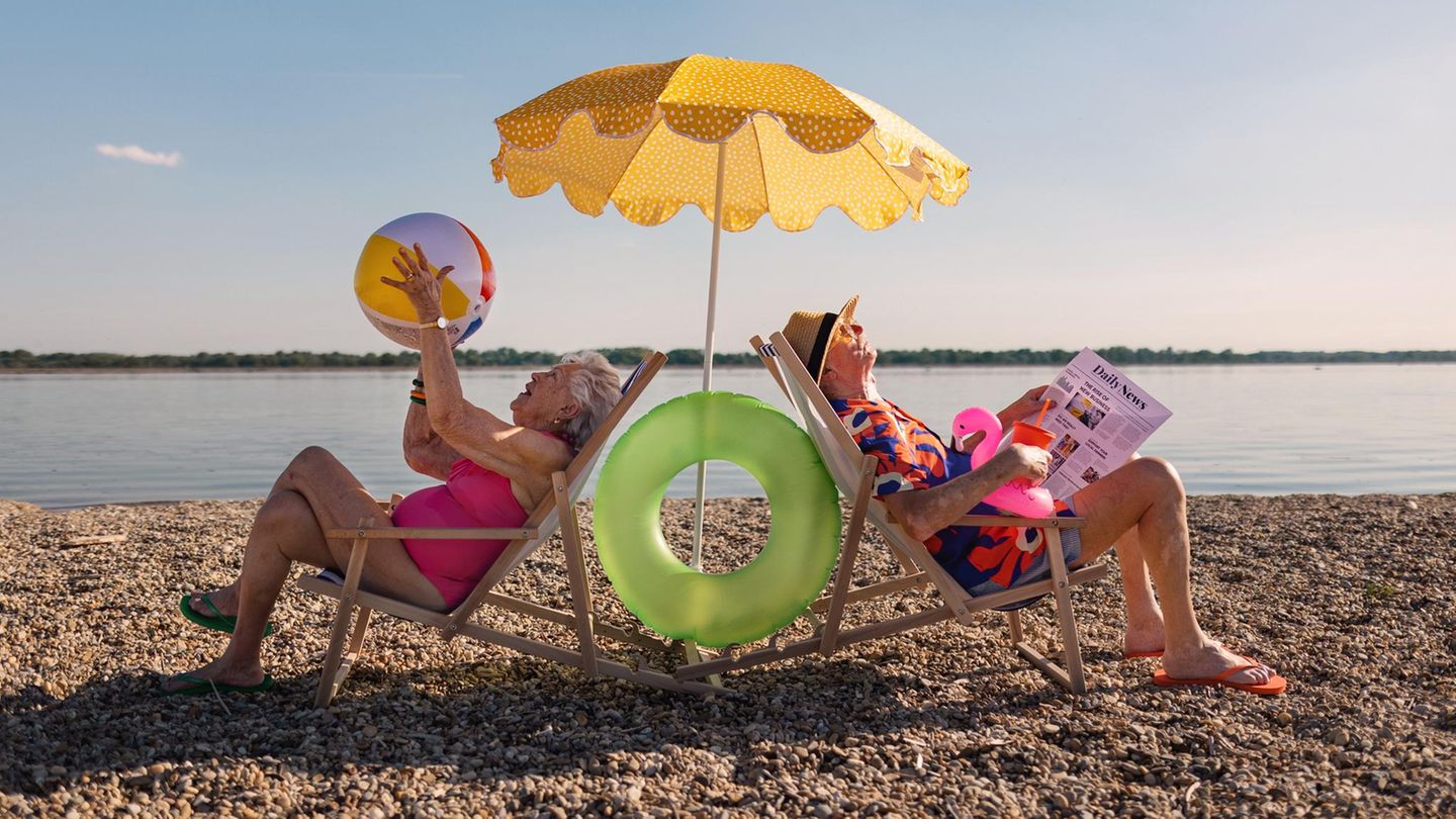 Porträt von Rentnern, die auf einer Strandliege unter einem Sonnenschirm sitzen.