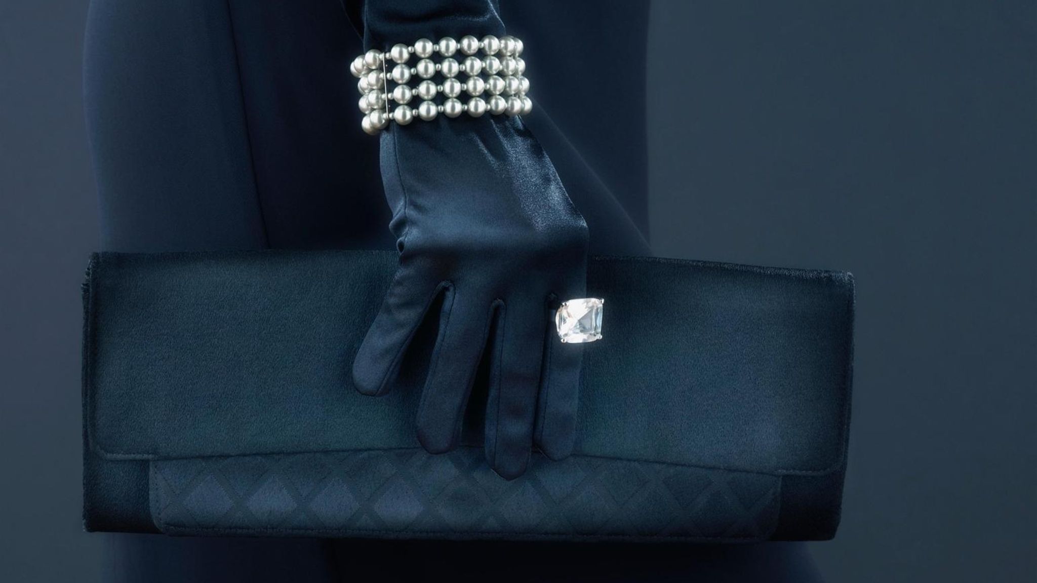 Frauenhand in einem schwarzem Lederhandschuh mit Perlenarmband, Diamantenring und schwarzer Handtasche
