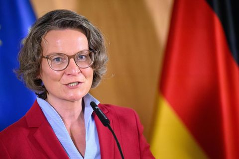 Ina Scharrenbach spricht vor einer Europa- und einer Deutschland-Flagge in ein Mikrofon und trägt dabei einen roten Blazer
