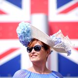 Eine Frau beim Royal Ascot vor einer Großbritannien-Flagge. Sie trägt einen Hut mit hellblauen Blumen und eine Sonnenbrille