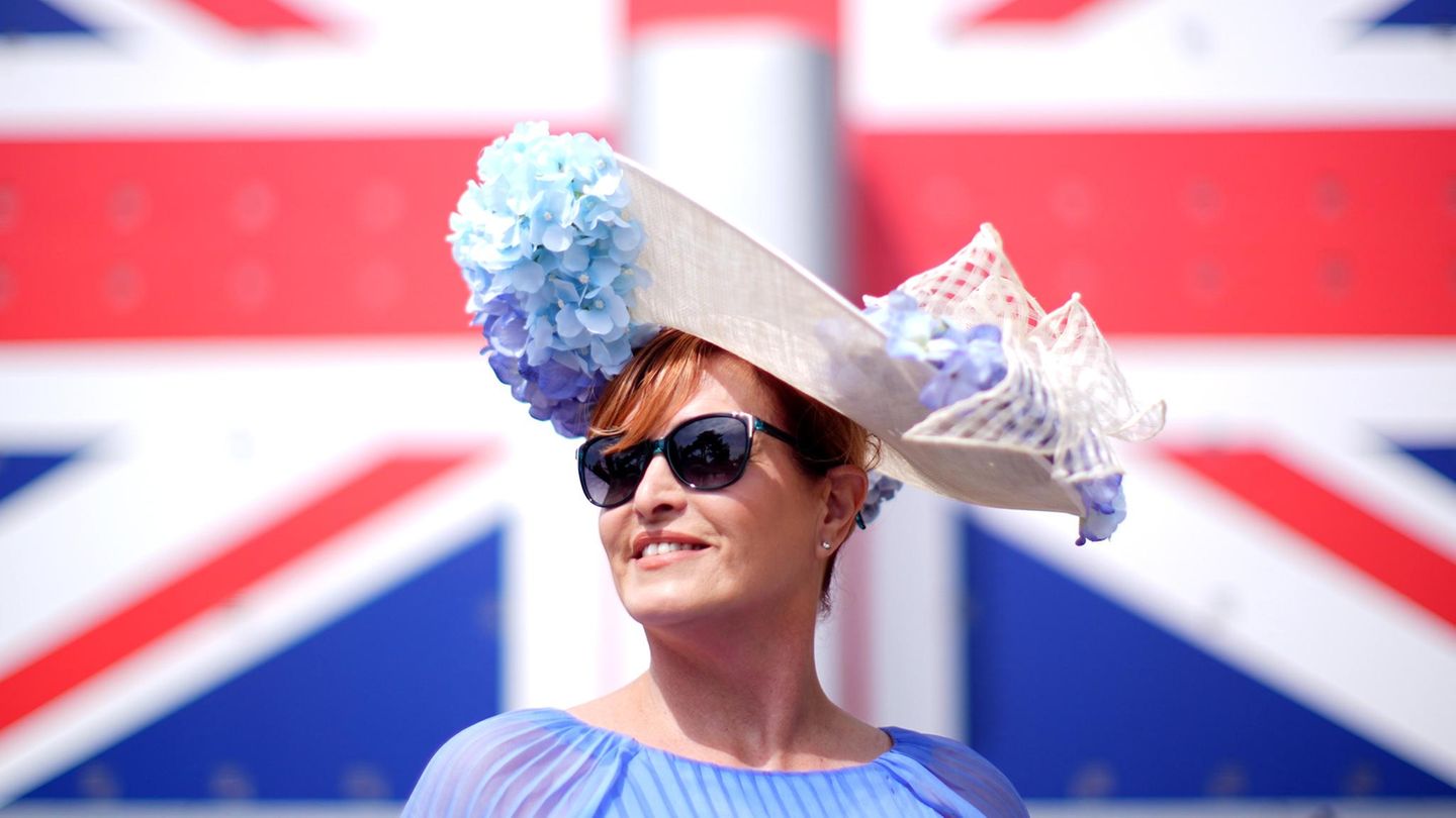 Eine Frau beim Royal Ascot vor einer Großbritannien-Flagge. Sie trägt einen Hut mit hellblauen Blumen und eine Sonnenbrille Eine Frau beim Royal Ascot vor einer Großbritannien-Flagge. Sie trägt einen Hut mit hellblauen Blumen und eine Sonnenbrille