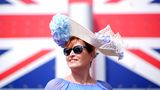 Eine Frau beim Royal Ascot vor einer Großbritannien-Flagge. Sie trägt einen Hut mit hellblauen Blumen und eine Sonnenbrille
