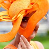 Eine Frau beim Royal Ascot mit einem blütenförmigen, orangefarbenen Hut. Sie schaut durch eines der Blütenblätter in die Kamera