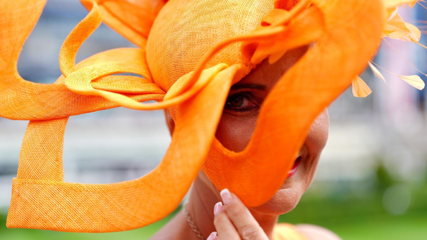 Eine Frau beim Royal Ascot mit einem blütenförmigen, orangefarbenen Hut. Sie schaut durch eines der Blütenblätter in die Kamera