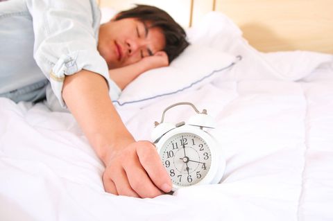 Ein Mann liegt schlafend auf dem Bett. Vor ihm befindet sich ein Wecker, dessen Zeiger auf 6 Uhr zeigen