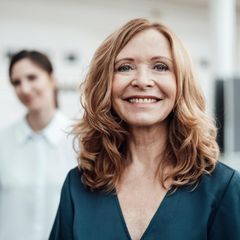 Eine Frau steht im Büro und schaut in die Kamera, hinter ihr eine Kollegin. Denkt sie schon an die Frührente?