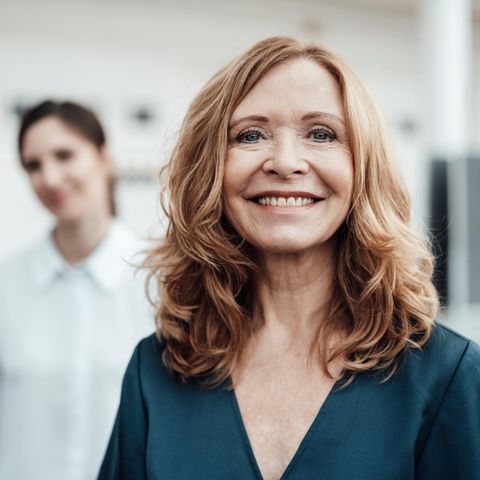 A woman stands in the office and looks into the camera, behind her a colleague. Does she think of the early retirement?