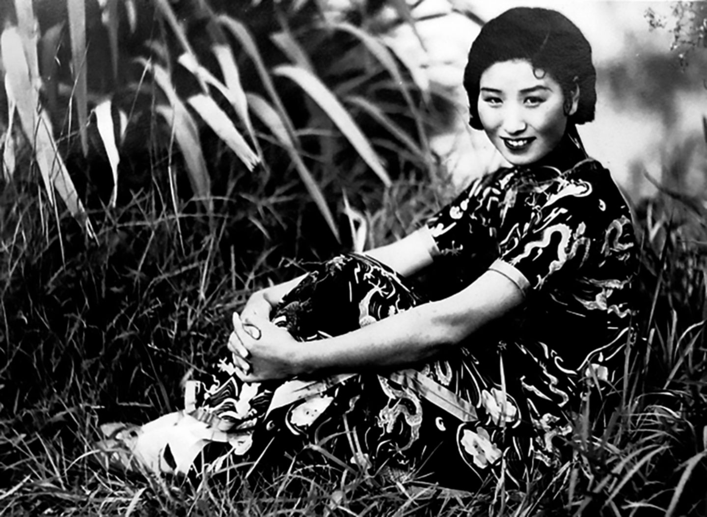 Eine Frau in einem chinesischen Kleid sitzt lächelnd im Gras, die Arme anmutig um die Beine geschlungen, schwarz-weiß, um 1940