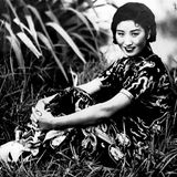 Eine Frau in einem chinesischen Kleid sitzt lächelnd im Gras, die Arme anmutig um die Beine geschlungen, schwarz-weiß, um 1940