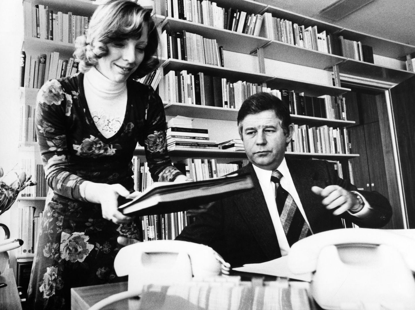 Eine schlanke Frau mit Akten in beiden Händen neben einem kurzhaarigen Mann an einem Schreibtisch, schwarz-weiß, 1970er Jahre