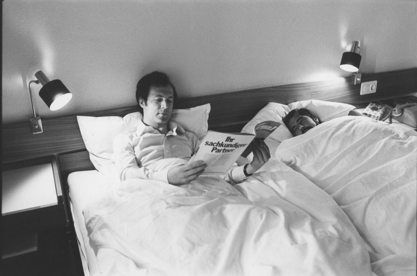 Beckenbauer und Gerd Müller liegen in einem Doppelbett, Beckenbauer liest ein Magazin