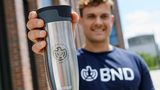 In einem Werbefoto hält jemand einen silbernen Kaffeebecher mit BND-Logo in die Kamera