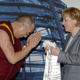 Angela Merkel hält einen weißen Seidenschal in den Händen, der Dalai Lama verbeugt sich.