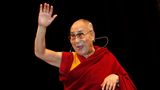 Der Dalai Dama winkt auf einer Bühne und schaut mit einem strahlenden Lächeln in die Menge