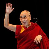 Der Dalai Dama winkt auf einer Bühne und schaut mit einem strahlenden Lächeln in die Menge
