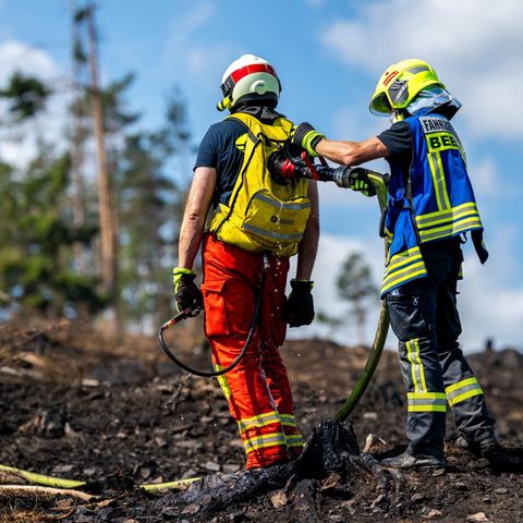 Zwei Feuerwehrleute stehen auf verkohltem Waldboden, während der eine mit einem Schlauch den Rucksack des anderen befüllt