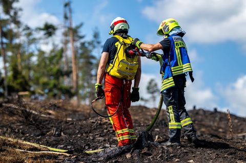 Zwei Feuerwehrleute stehen auf verkohltem Waldboden, während der eine mit einem Schlauch den Rucksack des anderen befüllt