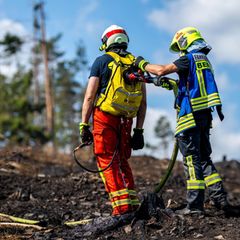 Zwei Feuerwehrleute stehen auf verkohltem Waldboden, während der eine mit einem Schlauch den Rucksack des anderen befüllt