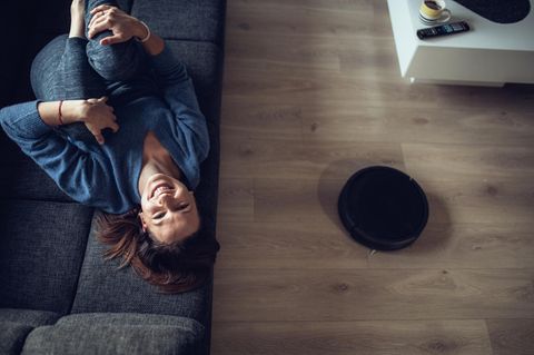 (Prime Day) Frau liegt lachend auf dem Sofa, auf dem Boden fährt ein Saugroboter.