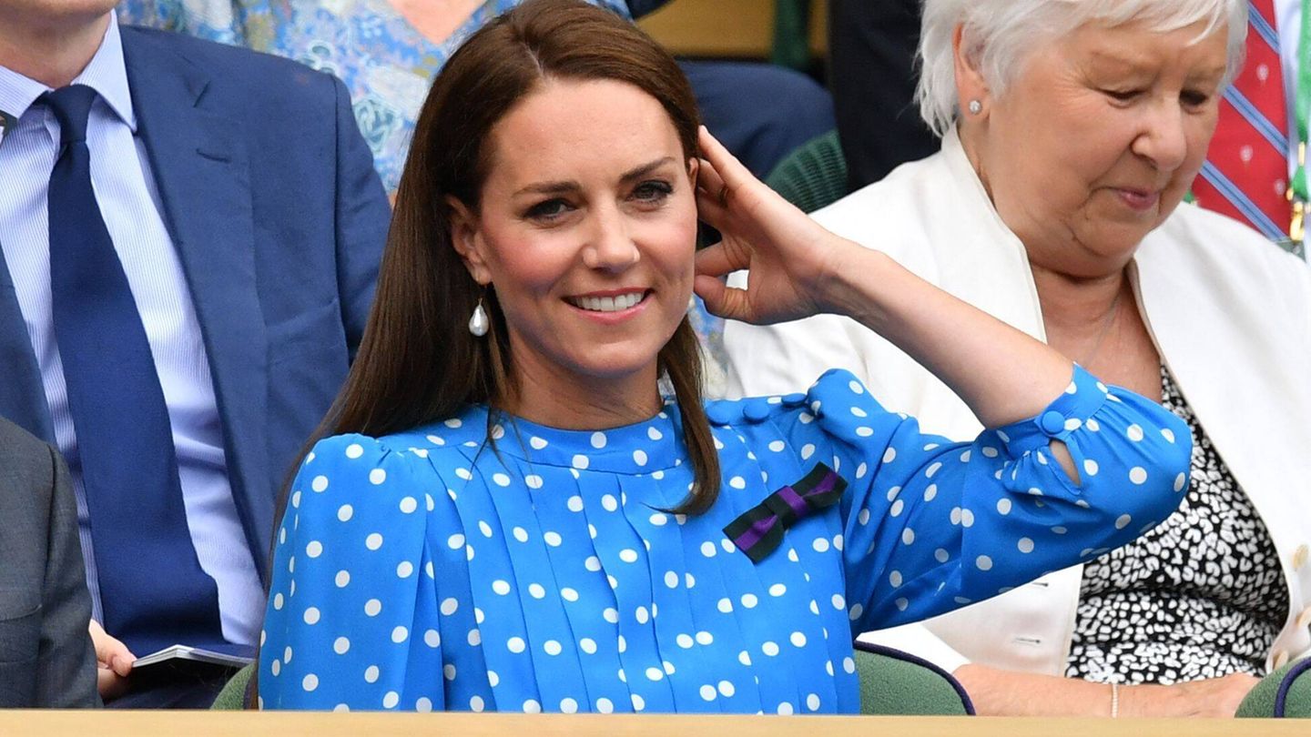 Kate in einem blauen Kleid mit weißen Punkten in der Logo, sie streicht ihr Haar zurück