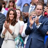 Kate klatscht auf der Tribüne, sie trägt ein eierschalenfarbenes Kleid