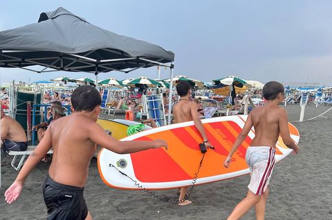 An einem Strand in Italien gehen drei Jungen mit einem SUP zum Wasser