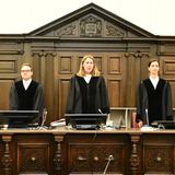 Zwei Frauen und ein Mann in schwarzen Roben stehen hinter der Richterbank im Block-Prozess, eingerahmt von